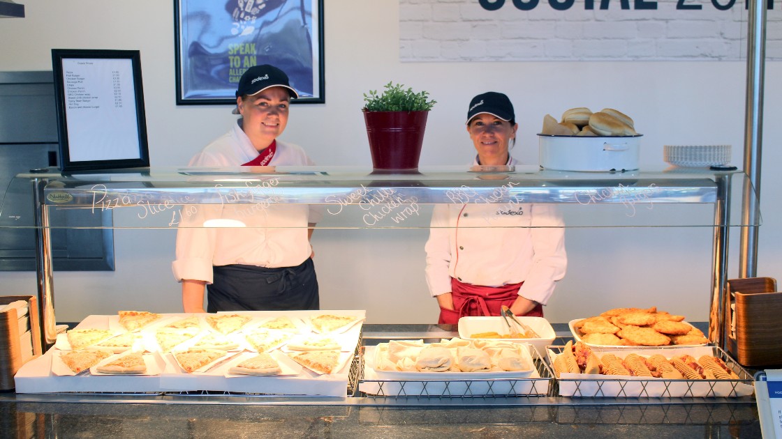 Two cafeteria staff behind a counter with food trays