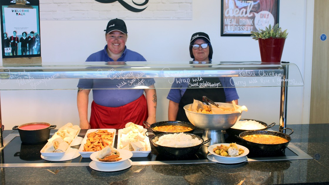 Two cafeteria staff behind a counter with various dishes on display