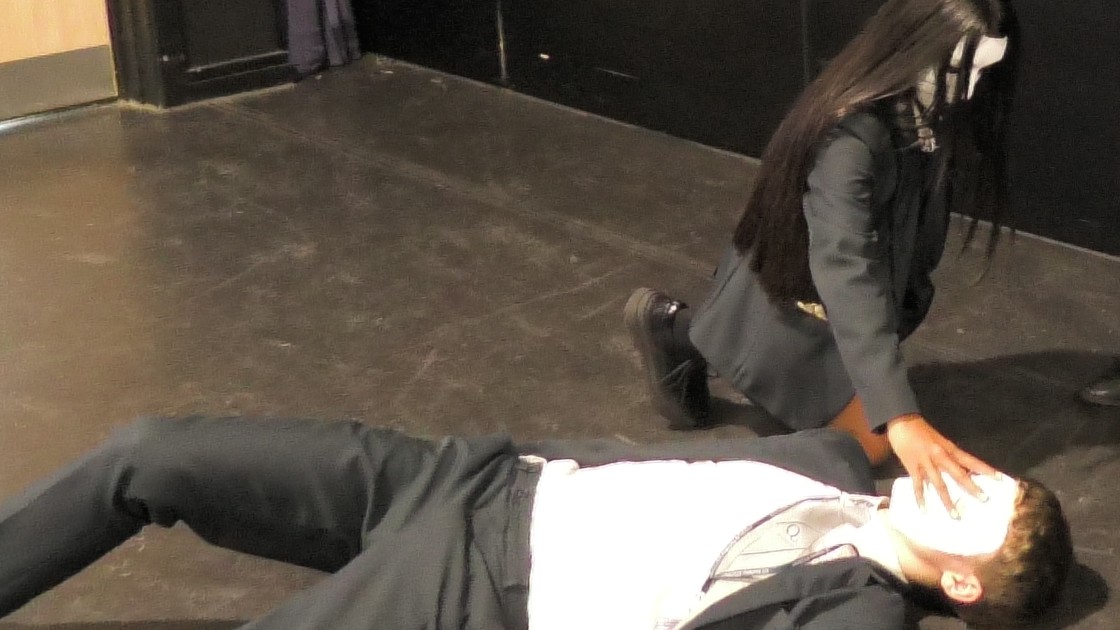 Two students in a drama lesson, one lying on the floor and the other wearing a mask, kneeling
