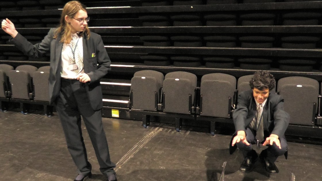 Two students in a drama lesson, one standing and gesturing, the other crouching on stage