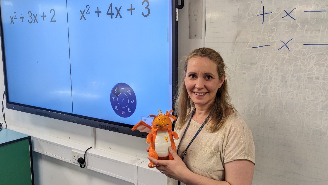 Teacher holding a dragon toy in front of a maths display