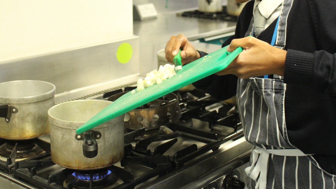 Student adding chopped vegetables to a pot on a stove