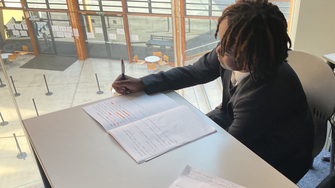 Student writing in a notebook at a desk near a window