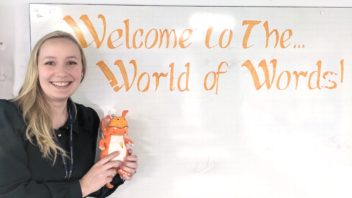 Teacher holding a small dragon toy next to a whiteboard with 'Welcome to The World of Words!' written on it