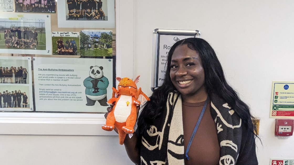 A teacher holding a small orange dragon toy, standing in front of a wall display about anti-bullying