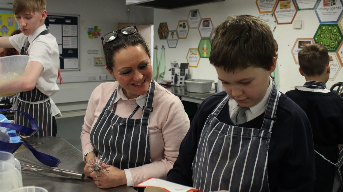 A teacher and a student wearing aprons in a kitchen setting, with the student holding a book