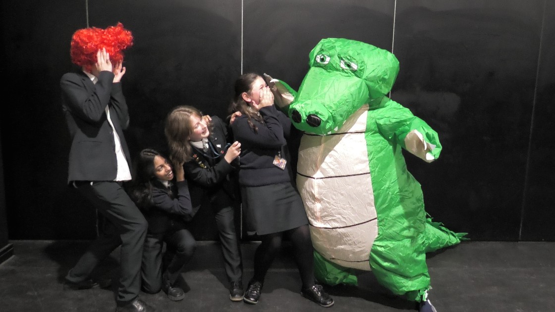 Students in costume, including a crocodile and a red wig, acting in a pantomime scene