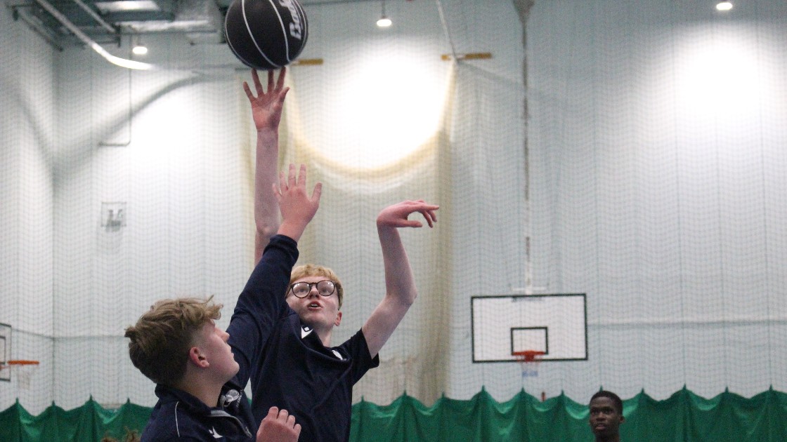 Two students playing basketball indoors, one jumping to shoot