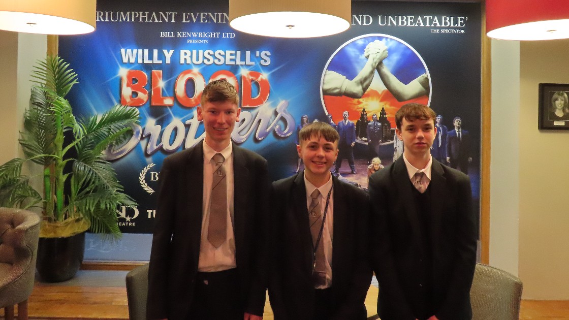 Three students in school uniforms standing in front of a Blood Brothers poster