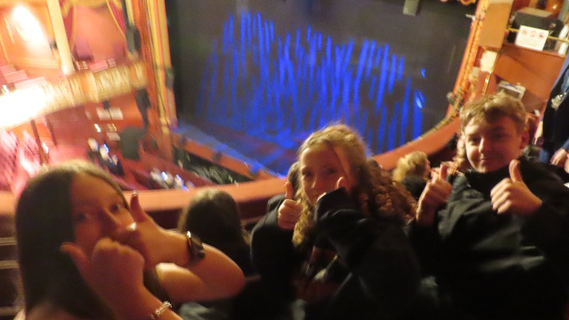 Students seated in a theatre, giving thumbs up, with a stage in the background