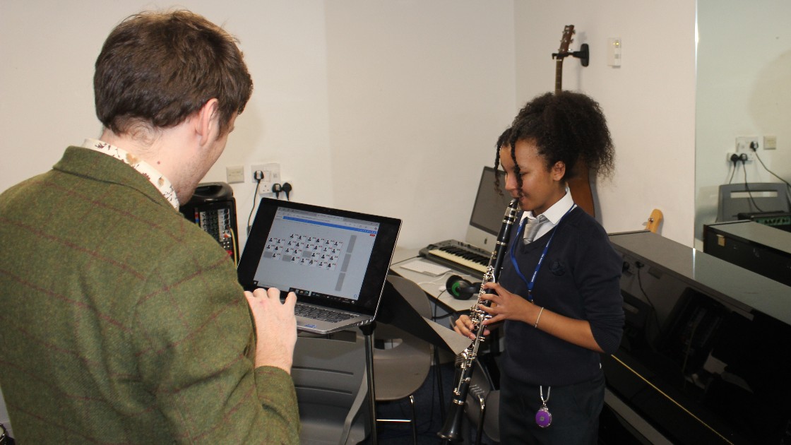 A teacher using a laptop while a student plays the clarinet in a music room