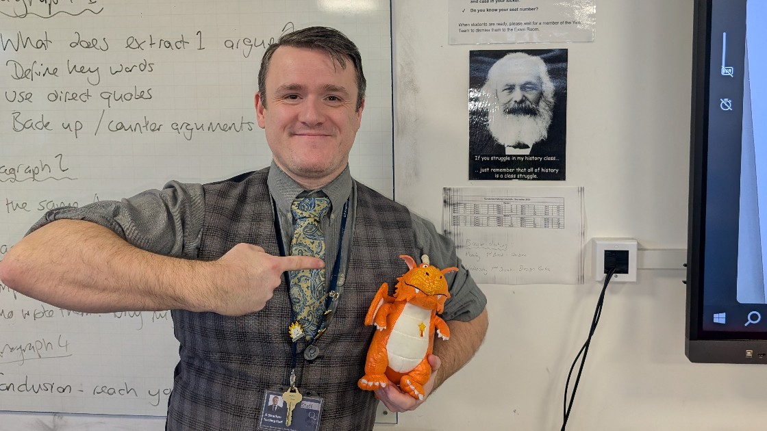 A teacher holding and pointing at an orange dragon toy in a classroom