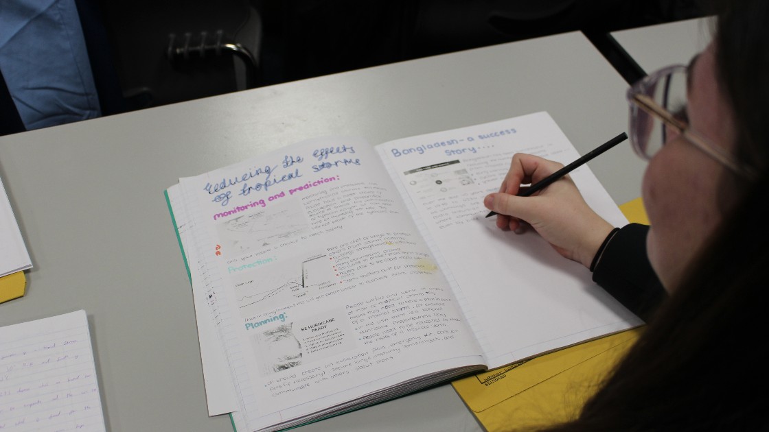 A student writing in a Geography exercise book with notes on Bangladesh