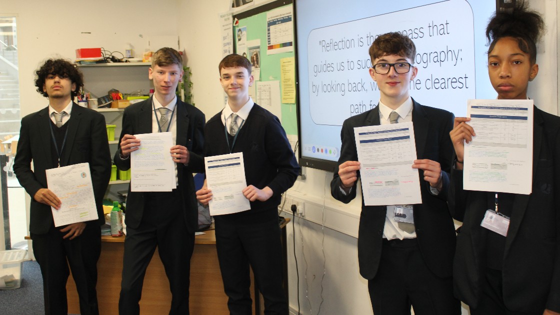 Students in a classroom holding up their completed Geography assignments