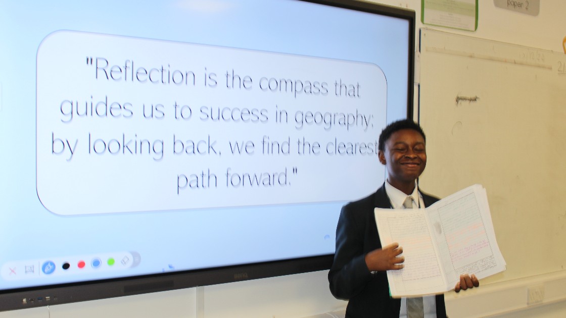 A student holding an exercise book stands in front of a screen displaying a quote about reflection in geography