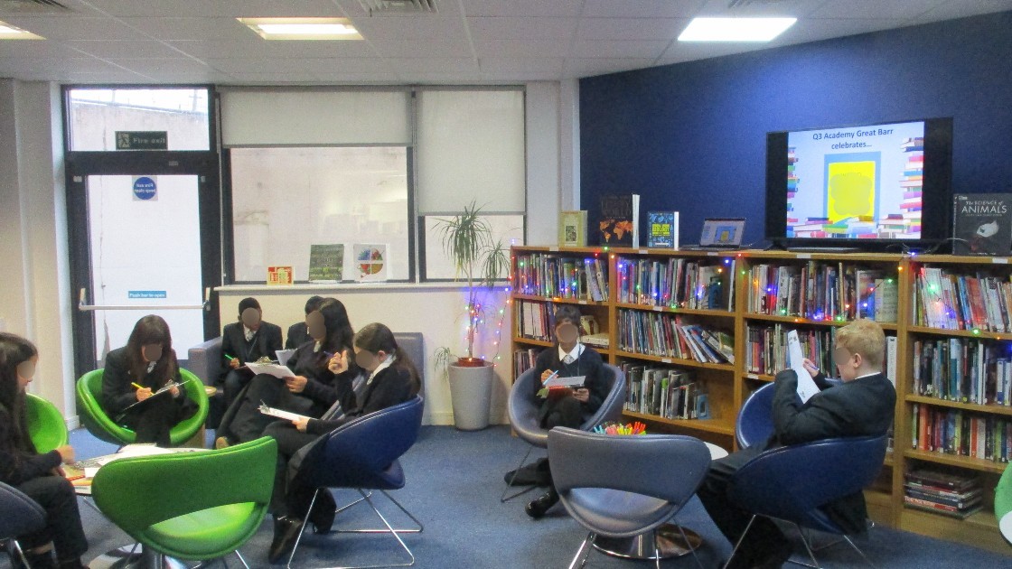 Students sitting in a library reading books, with bookshelves and a screen in the background