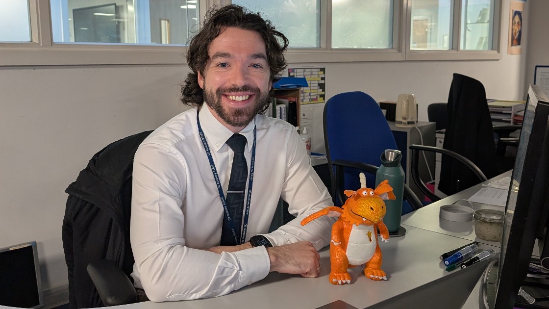 A teacher sitting at a desk with a small orange dragon model