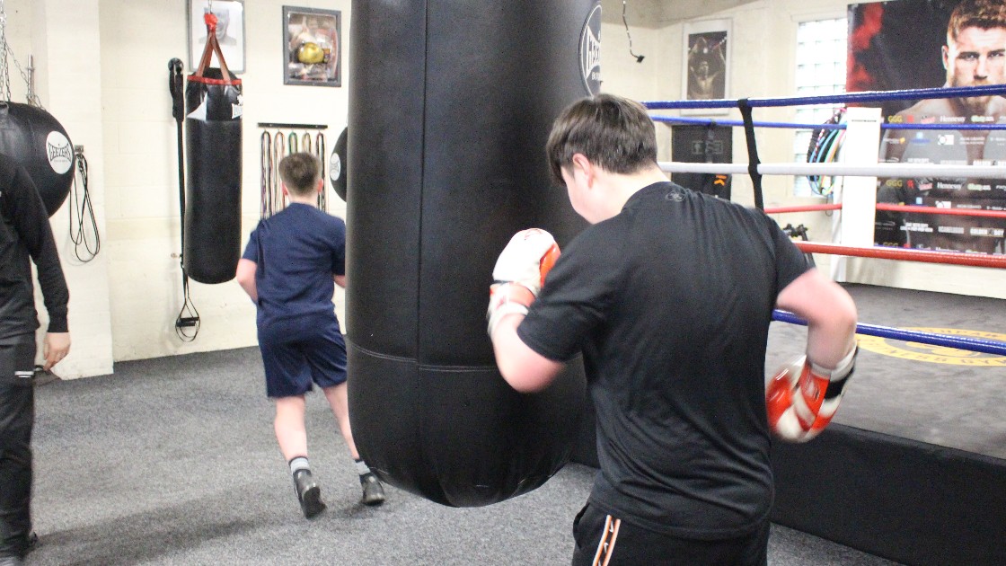 Two students in a boxing gym, one punching a heavy bag and the other running