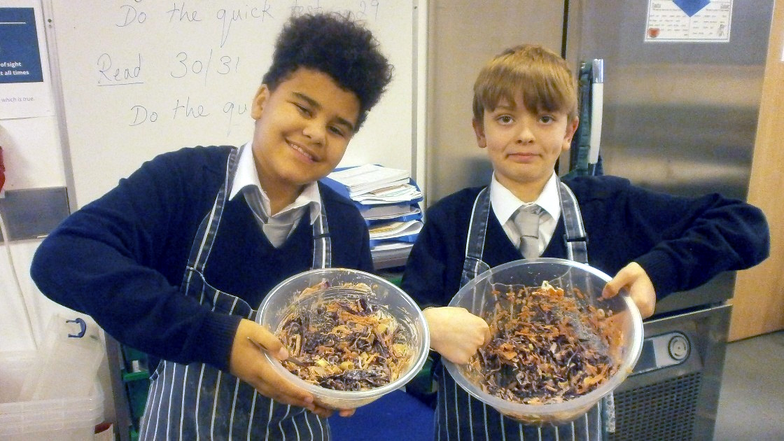 Two students in aprons holding bowls of mixed salad ingredients in a classroom