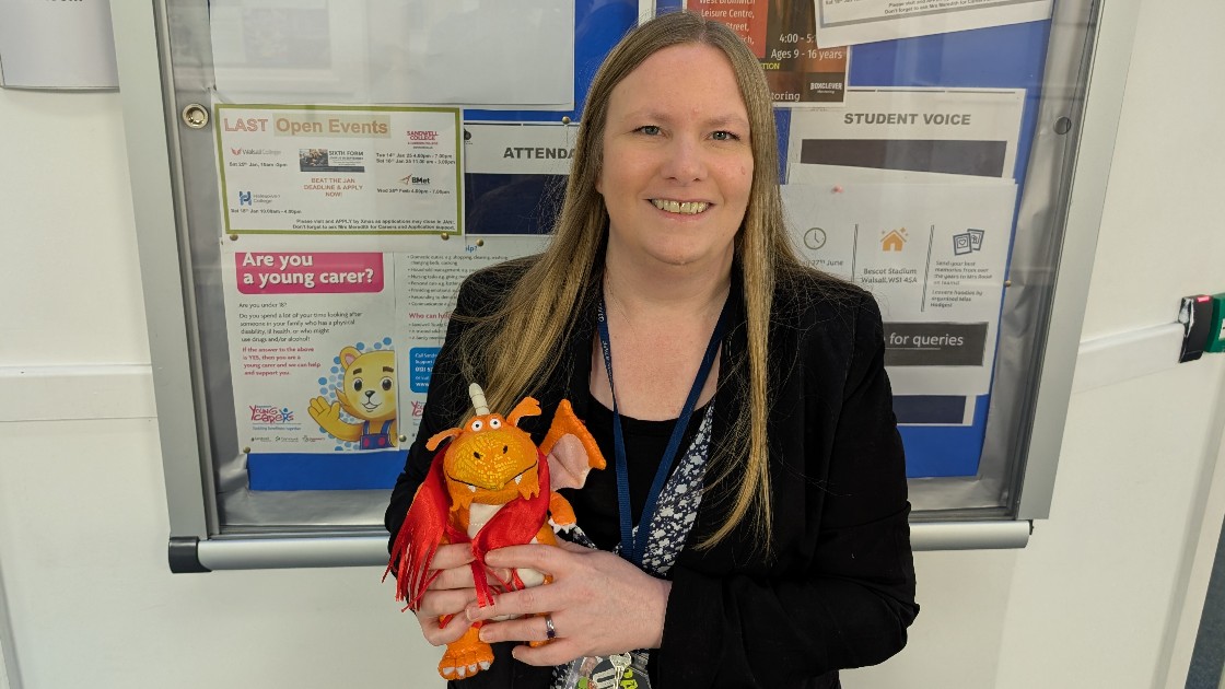 A teacher holding a small dragon toy, standing in front of a noticeboard