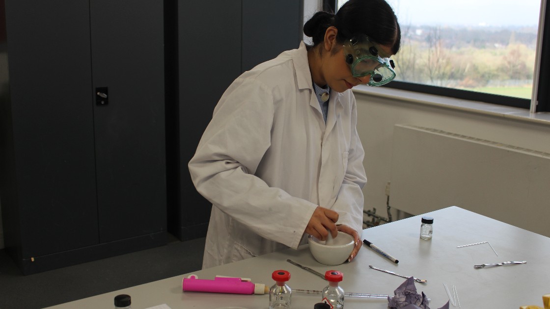 A student wearing safety goggles and a lab coat working with a mortar and pestle in a science classroom