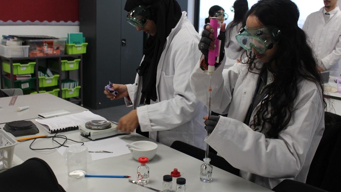 Students in a science lab wearing goggles and lab coats, using pipettes and scales