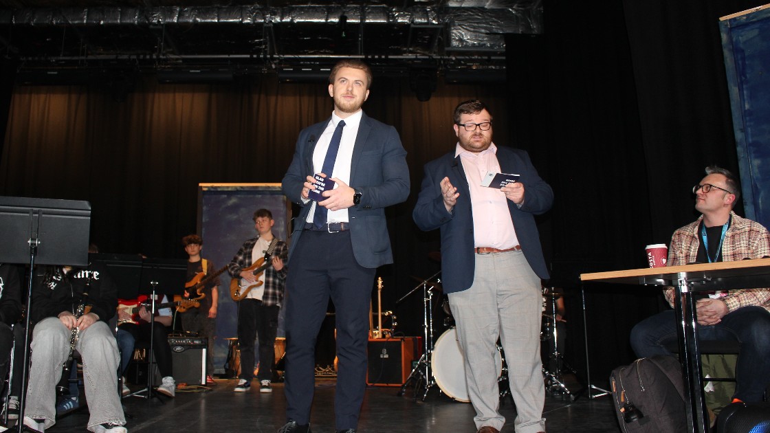Two teachers on stage speaking, with a student band performing in the background