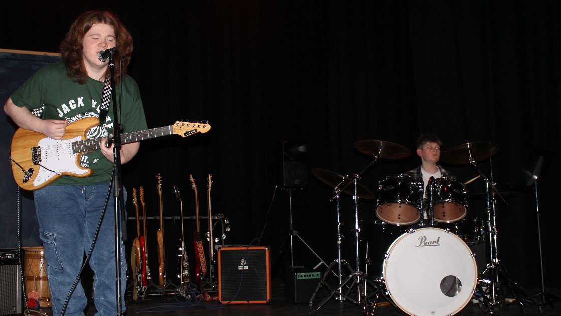 A student playing guitar and singing, with another student playing drums on stage