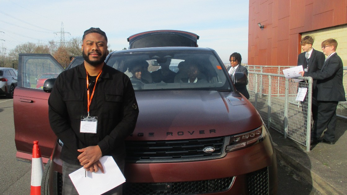 A professional stands in front of a Range Rover with students visible inside and nearby, holding papers