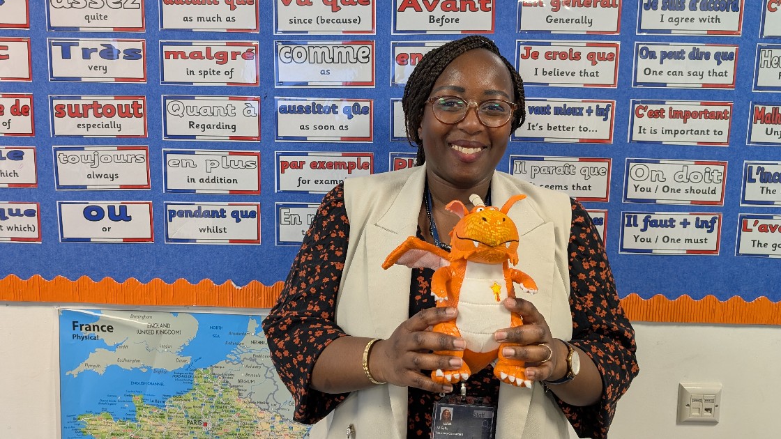A teacher holding a small orange dragon toy in a classroom with French language posters on the wall