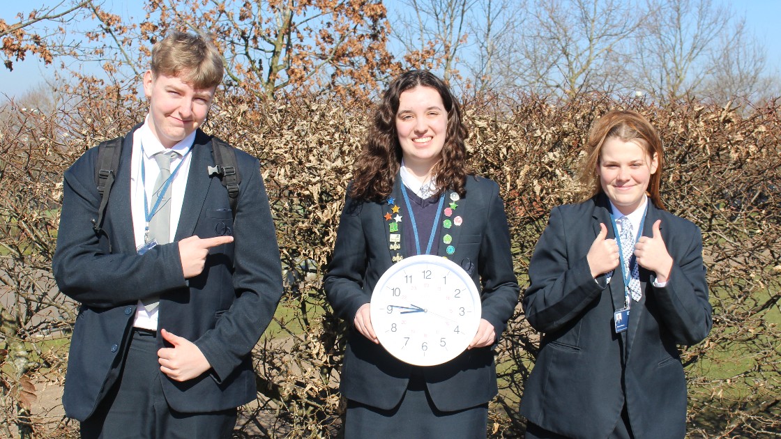 Three students in uniform outdoors, one holding a clock