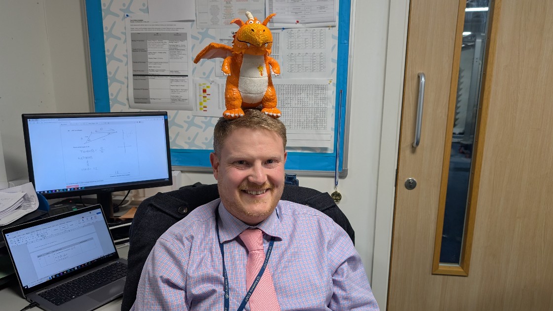 A teacher with a toy dragon on their head, sitting in an office