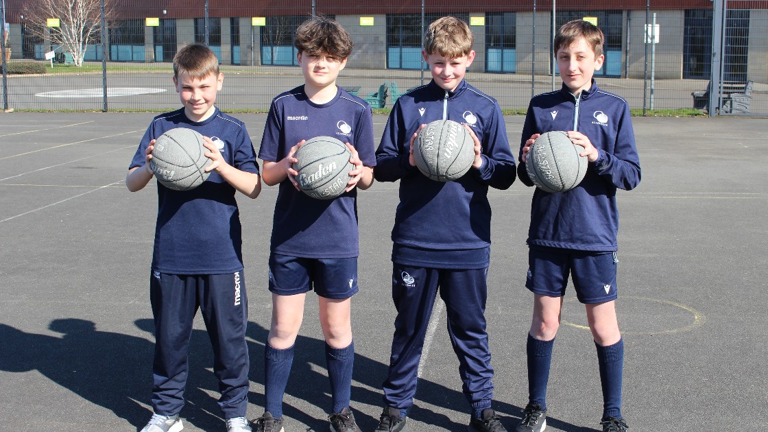 Four students in sports kit holding basketballs on an outdoor court