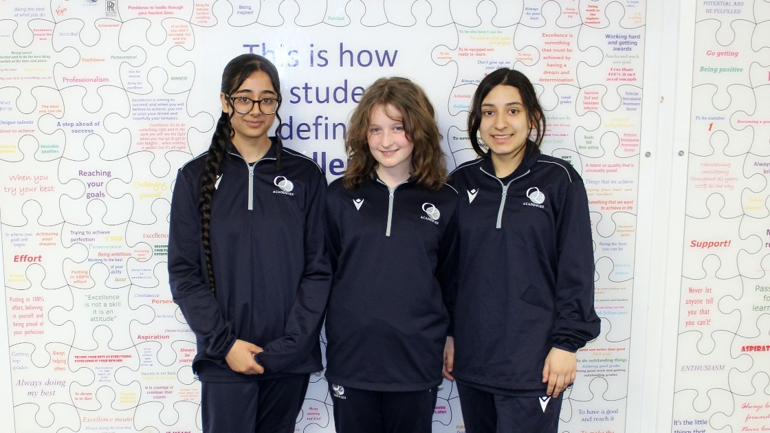 Three students wearing matching sports kits stand in front of a wall with motivational text