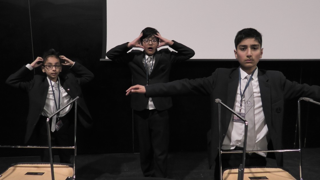 Three students in school uniforms performing a dramatic scene on stage, with expressive gestures and props