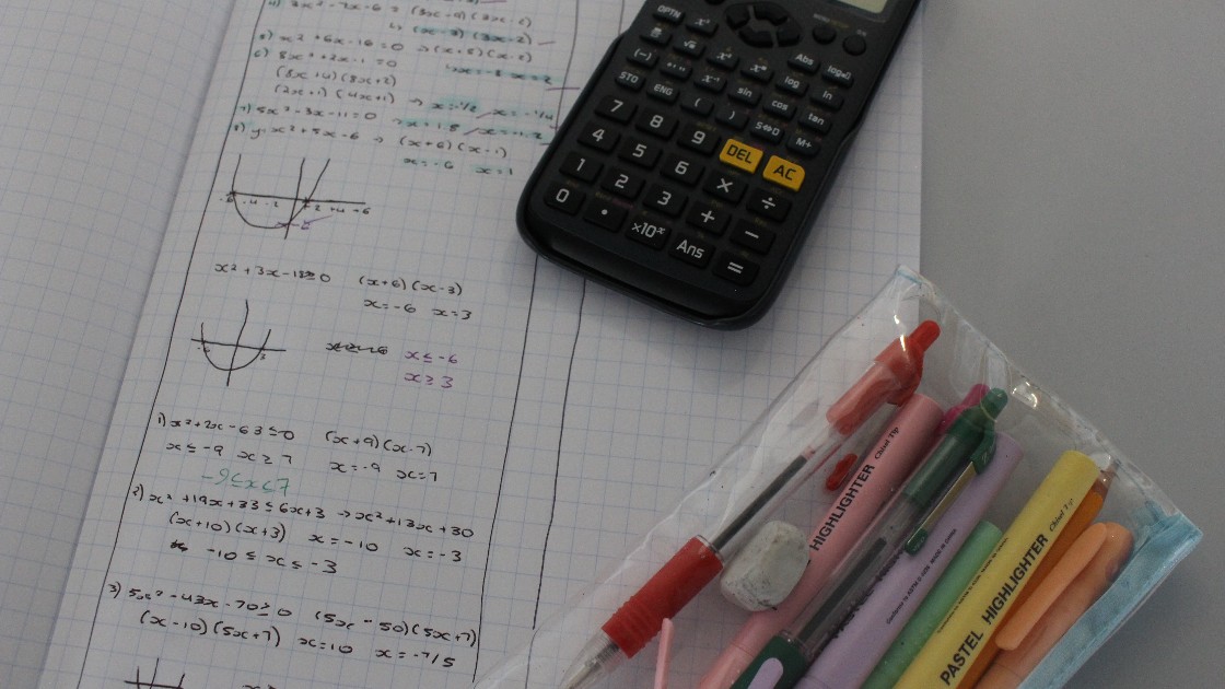Maths notes with equations and graphs, a calculator, and stationery on a desk