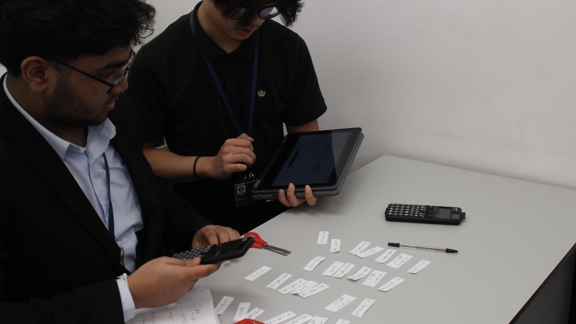 Two students working on a maths activity with calculators, a tablet, and small paper cards on a desk