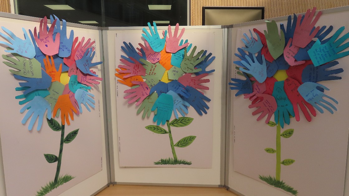 Three colourful paper flowers made of hand-shaped cutouts with messages written on them, displayed on boards.