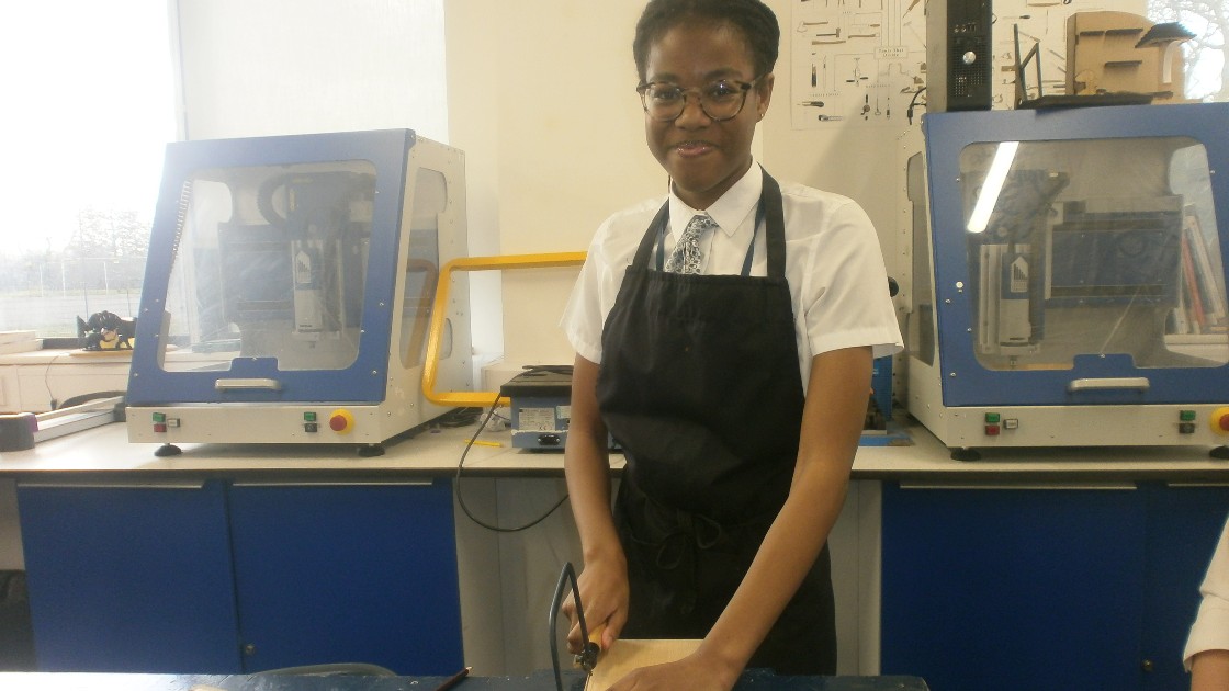 A student wearing an apron works on a project in a design and technology classroom, with machinery in the background