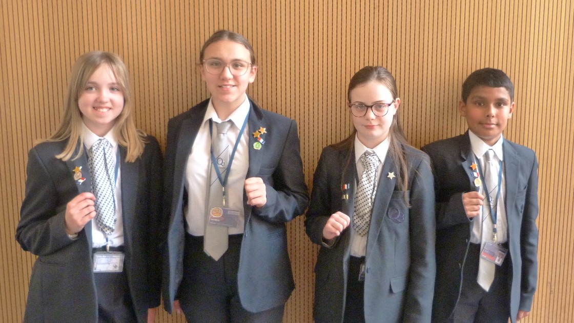 Four students in uniform holding up badges or awards, standing in front of a wooden wall