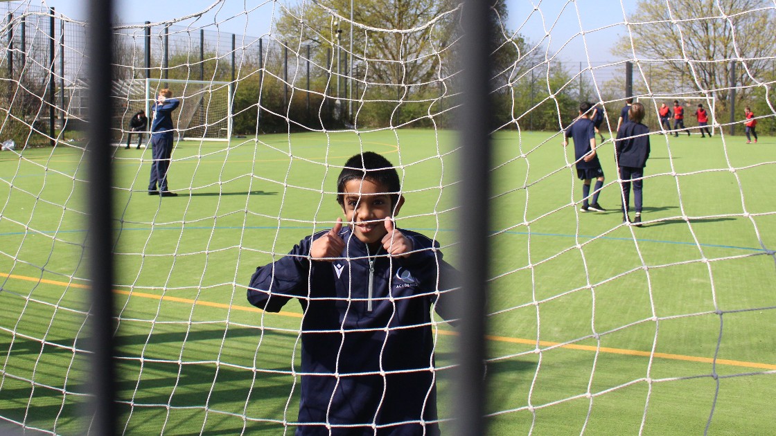 A student smiling and posing behind a football net on a school field during a PE lesson