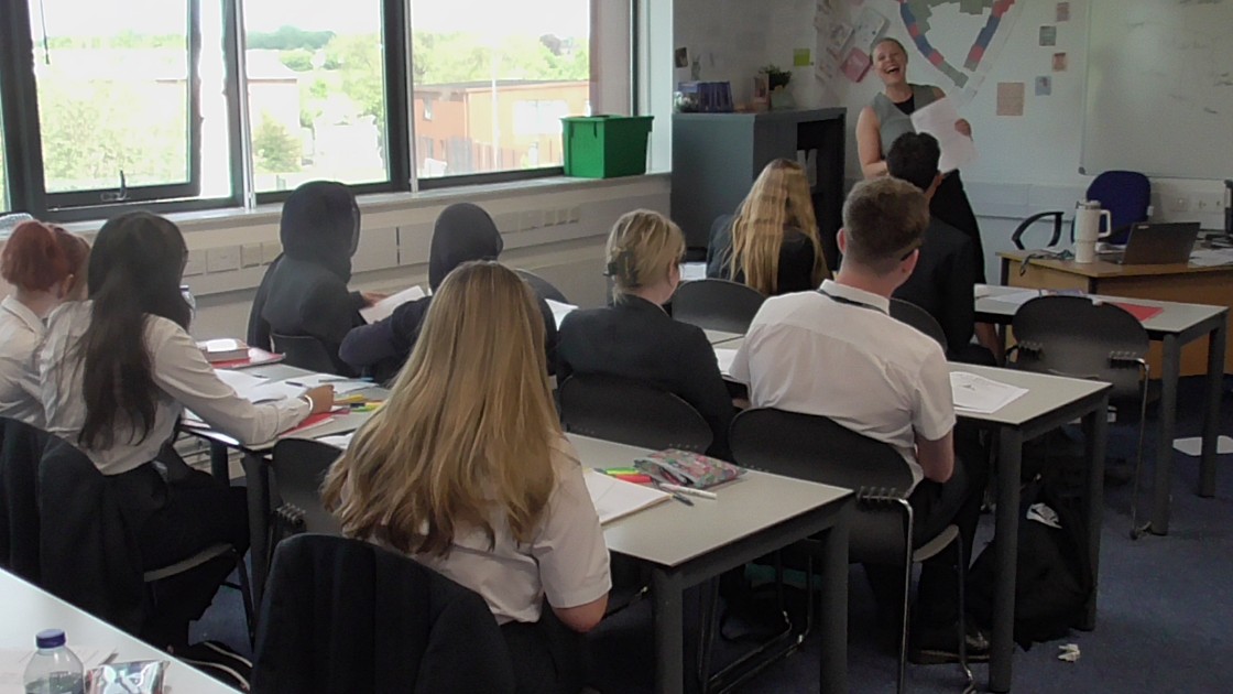 A teacher stands at the front of a classroom, speaking to students seated at desks during an English lesson