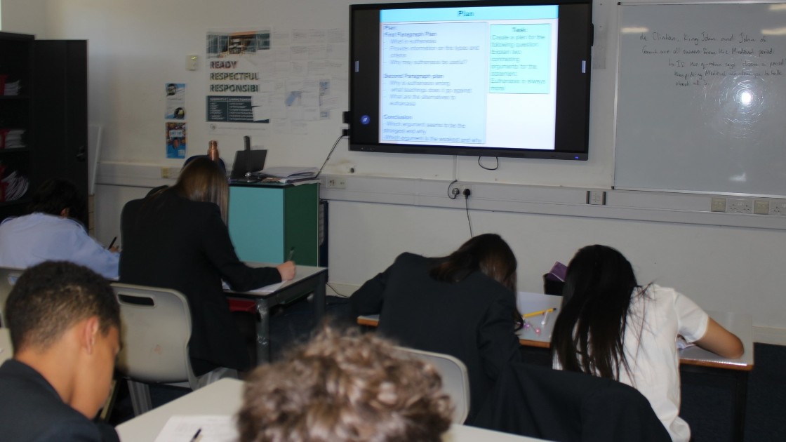 Students sitting at desks in a classroom, writing while a lesson plan is displayed on the board