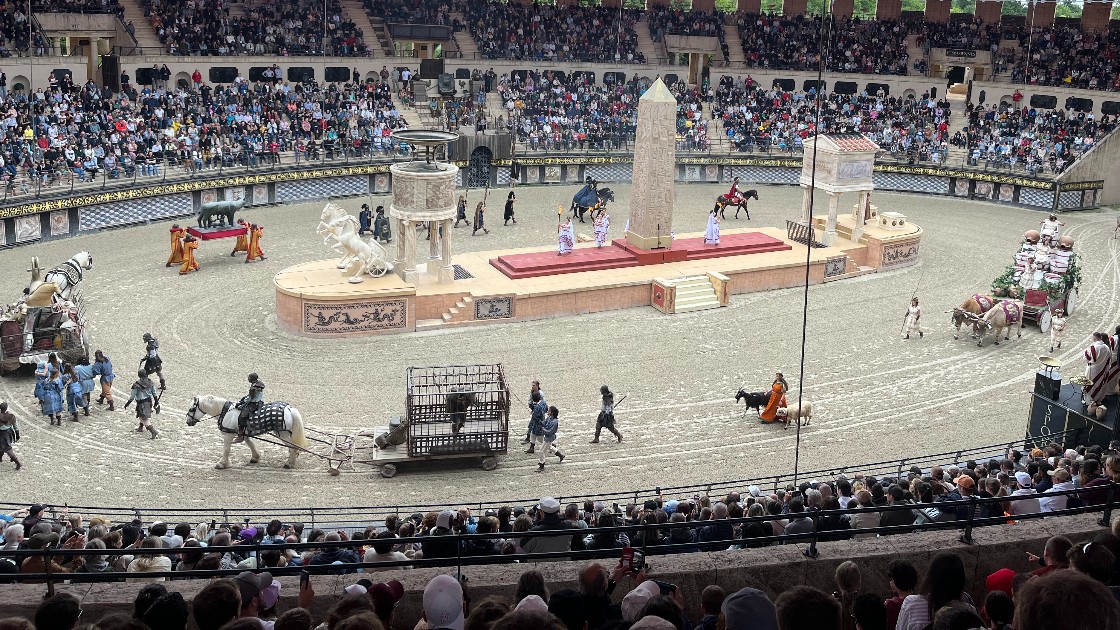 A large arena with a crowd watching a historical reenactment featuring actors in Roman costumes, horses, and chariots