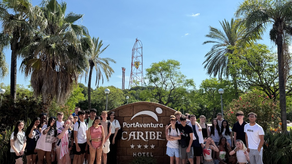 A group of students posing in front of the PortAventura Caribe Hotel sign with palm trees and Red Force in view.