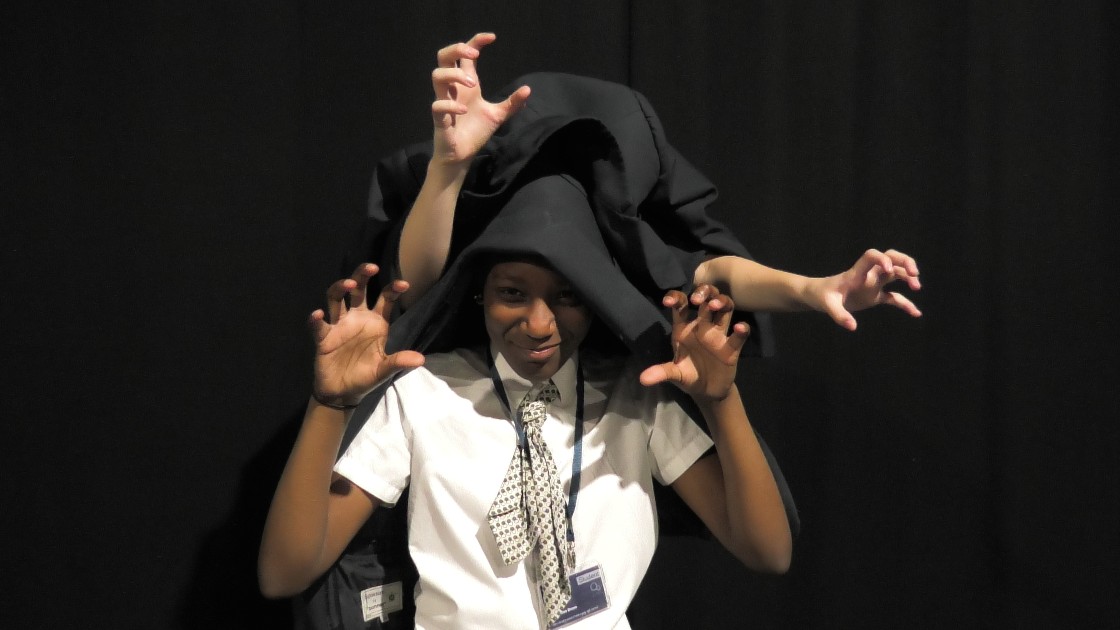 Two students performing a drama activity, with one student in front and another behind, both making dramatic hand gestures. The student in front is wearing a tie and has a jacket draped over their head