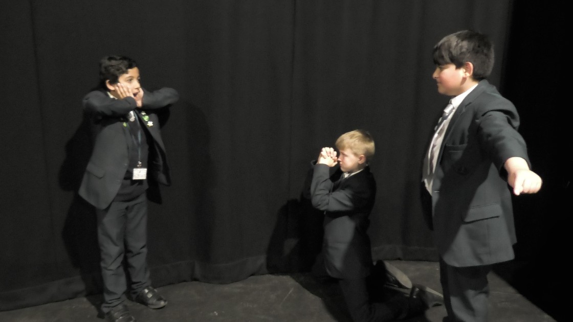Three students in school uniform acting out a scene in front of a black curtain. One student stands with arms outstretched, another kneels with hands clasped, and the third stands with hands near their face