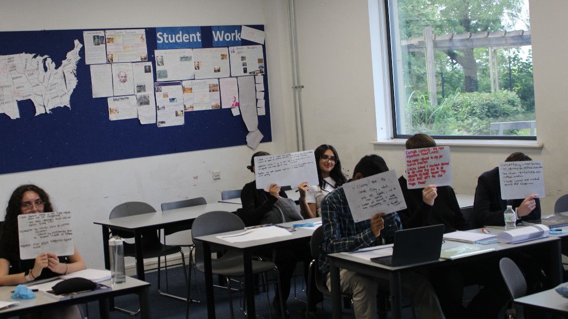 Students in a classroom holding up whiteboards with written answers, with a display of student work on the wall