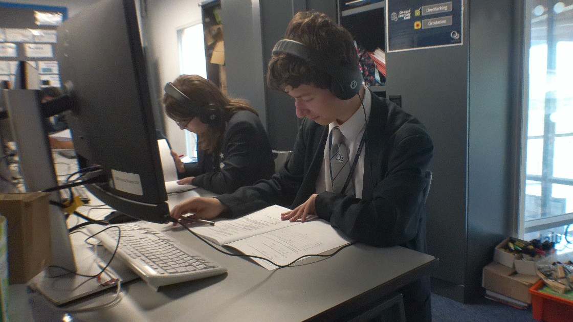 Two students wearing headphones working at computers with worksheets in a classroom