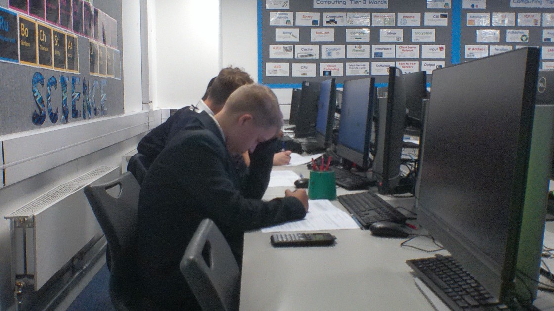 Students working at computers in a classroom, writing on paper